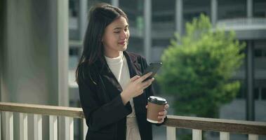 imágenes de joven asiático mujer de negocios en traje participación un taza de café y utilizando un móvil teléfono mientras en pie en un moderno negocio edificio. negocio y personas conceptos. video