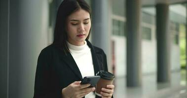 Aufnahmen von jung elegant asiatisch beschäftigt Geschäft Frau im ein passen halten Handy, Mobiltelefon Telefon und ein Tasse von Kaffee während Stehen im Vorderseite von ein modern Geschäft Gebäude. Geschäft und Menschen Konzepte. video