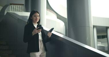 imágenes de joven asiático mujer de negocios en traje utilizando un teléfono inteligente y ordenador portátil mientras caminando abajo en un moderno negocio edificio. negocio y personas conceptos. video