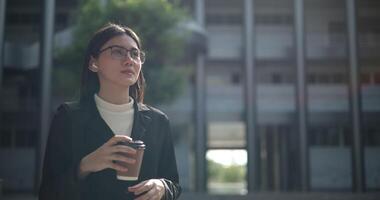 Footage of Young smiling elegant Asian busy businesswoman wearing glasses in a suit drinking coffee while standing in front of a modern business building. Business and people concepts. video