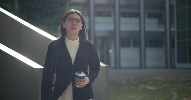 imágenes de joven elegante asiático ocupado mujer de negocios vistiendo lentes en un traje participación un taza de café mientras caminando en frente de un moderno negocio edificio. negocio y personas conceptos. video