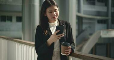 Aufnahmen von jung asiatisch Geschäftsfrau im passen halten ein Tasse von Kaffee und mit ein Handy, Mobiltelefon Telefon während Stehen im ein modern Geschäft Gebäude. Geschäft und Menschen Konzepte. video