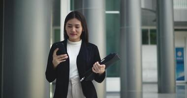 främre se skott, antal fot av ung leende elegant asiatisk upptagen företag kvinna i en kostym använder sig av en smartphone och gående i främre av en modern företag byggnad. företag och människor begrepp. video