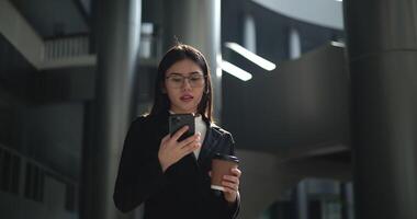 beeldmateriaal van jong Aziatisch bezig zakenvrouw vervelend bril in een pak Holding een kop van koffie en pratend Aan de telefoon terwijl staand in een modern bedrijf gebouw. bedrijf en mensen concepten. video