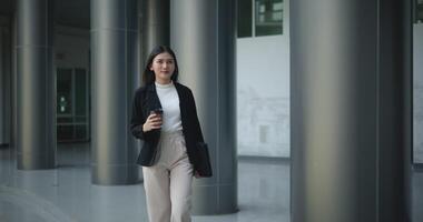 beeldmateriaal van jong glimlachen elegant Aziatisch bezig bedrijf vrouw in een pak Holding een kop van koffie en wandelen in voorkant van een modern bedrijf gebouw. bedrijf en mensen concepten. video