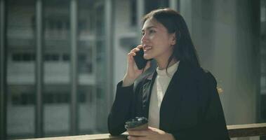Aufnahmen von jung asiatisch Geschäftsfrau im passen halten ein Tasse von Kaffee und reden auf ein Handy, Mobiltelefon Telefon während Stehen im ein modern Geschäft Gebäude. Geschäft und Menschen Konzepte. video