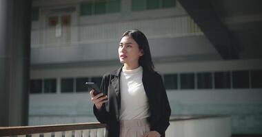 beeldmateriaal van gelukkig jong Aziatisch zakenvrouw in pak gebruik makend van een smartphone terwijl wandelen in een modern bedrijf gebouw. bedrijf en mensen concepten. video