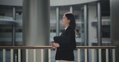 lado ver disparo, imágenes de contento joven asiático mujer de negocios en traje participación un ordenador portátil y taza mientras caminando en un moderno negocio edificio. negocio y personas conceptos. video