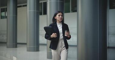 Footage of Young Asian businesswoman in suit holding a laptop and cup and running in a hurry in a modern business building. Business and people concepts. video