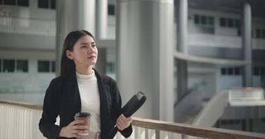 metraggio di contento giovane asiatico donna d'affari nel completo da uomo Tenere un' il computer portatile e tazza mentre a piedi nel un' moderno attività commerciale costruzione. attività commerciale e persone concetti. video