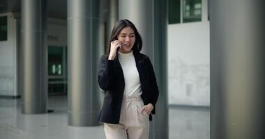 Front view shot, Footage of Young smiling elegant Asian busy business woman in a suit talking with a smartphone and walking in front of a modern business building. Business and people concepts. video