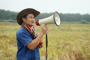 Asian man farmer wears hat, holds megaphone at agriculture field. Concept, communication , announce. Protest. Protester. Bullhorn public address. Loudspeaker technology for outdoor announcement. photo