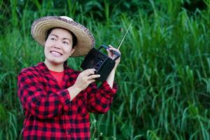 Asian woman farmer, holds AM, FM transistor radio receiver on shoulder. Concept  Happy working along with music, news, information ,advertisement from radio. Country lifestyle. Analog technology. photo