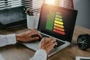 persona mano utilizando ordenador portátil computadora con casa icono energía eficiencia escala imagen en oficina escritorio, concepto de ecológico y bio energético casa. energía clase.. foto