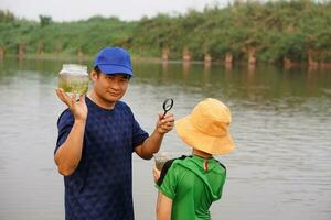 Asian man and boy are at lake, The man holds jar of sample water and magnifying glass. Concept, ecology exploring. Education. Learning by doing. Life experience photo