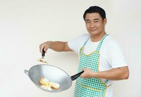Handsome Asian man is cooking fried eggs, wears white shirt and apron, holds frying pan and ladle spatula for frying. Concept, love cooking. Kitchen lifestyle. Look at camera. Copy space for text photo