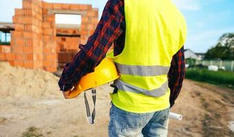 Construction worker on site zone. Worker in yellow vest holding hard hat and staying in front of the building zone. Construction worker on the work zone. photo