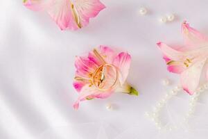 Two gold engagement rings inside a lovely pink astromeria flower on a white satin background with pearls. Wedding background. A copy space. photo