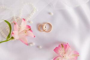 Two gold wedding rings lie on a white satin background with a scattering of pearls and pink astromeria flowers. Wedding background. Top view. photo