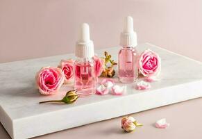 Two cosmetic bottles with a pipette and a white cap with a cosmetic product based on rose oil on a white marble podium with flowers. photo