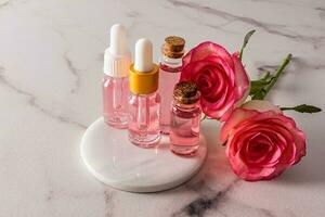 cosmético Rosa petróleo y Rosa agua en varios vaso botellas en un blanco redondo mármol podio con rosas. natural productos cosméticos. adicional cuidado. foto