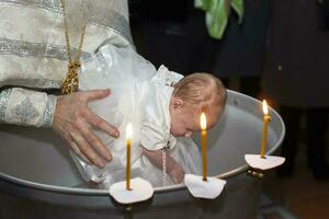 ortodoxo rito de bautismo de un niño. el manos de el sacerdote bañarse el bebé en el bautismal baño. foto