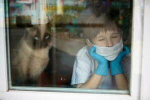 Quarantine from a child of COVID-19.A boy in a mask and medical gloves outside the window. photo