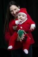 mamá y pequeño niño a Navidad. un mujer sostiene su hijo en su brazos, quien es vestido en Papa Noel claus ropa. foto