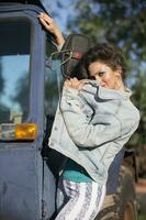 hermosa joven mujer en un mezclilla chaqueta posando en contra el fondo de un tractor. niña en el estilo de el años ochenta foto