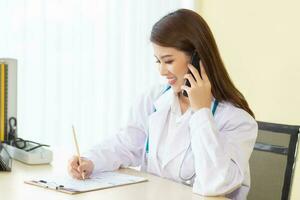 Professional Young Asian woman doctor wearing a white robe and stethoscope sitting with use smartphones to talk inquire follow up on patients symptoms in the examination room at hospital. photo