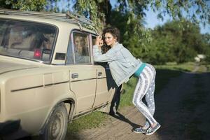 Funny young woman in a denim jacket posing amusingly against the background of an old car in the village. photo