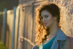 A beautiful girl in a denim jacket with bright makeup stands near a wooden fence illuminated by the evening sun. photo