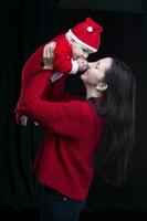 mamá y pequeño niño a Navidad. un mujer sostiene su hijo en su brazos, quien es vestido en Papa Noel claus ropa. foto