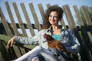 A beautiful young woman in a denim jacket with bright makeup sits with a chicken on the background of an old fence. photo