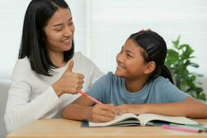 madre enseñando lección para hija. asiático joven pequeño niña aprender a hogar. hacer deberes con tipo madre ayuda, animar para examen.. niña contento educación en el hogar mamá Consejo educación juntos. foto