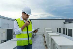 Asian maintenance engineer works on the roof of factory. contractor inspect compressor system and plans installation of air condition systems in construction. technology, online, mobile application. photo