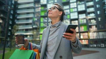Frau Gehen auf ein Geschäft Kreis hält wegbringen Kaffee Einkaufen Taschen und Verwendet Smartphone video