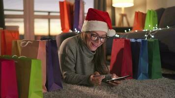 Happy woman with glasses wearing a santa claus hat is lying on the carpet and makes an online purchase using a credit card video