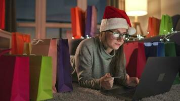 contento mujer con lentes vistiendo un Papa Noel claus sombrero es acostado en el alfombra y hace un en línea compra utilizando un crédito tarjeta video