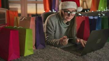 Happy woman with glasses wearing a santa claus hat is lying on the carpet and makes an online purchase using a credit card video