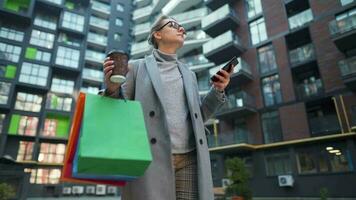 mujer caminando en un negocio distrito sostiene para llevar café compras pantalones y usos teléfono inteligente video