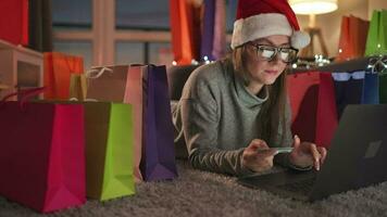 Happy woman with glasses wearing a santa claus hat is lying on the carpet and makes an online purchase using a credit card video