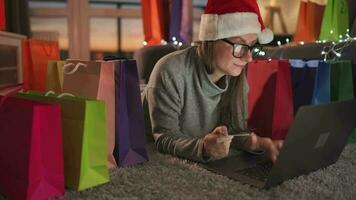 Happy woman with glasses wearing a santa claus hat is lying on the carpet and makes an online purchase using a credit card video