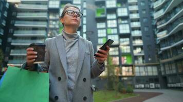 Woman Walking on a Business District Holds Takeaway Coffee Shopping Bags and Uses Smartphone video