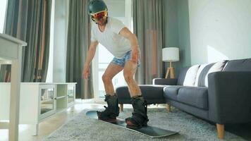 Fun video. Man in shorts and a T-shirt depicts snowboarding on a carpet in a cozy room. Waiting for a snowy winter video