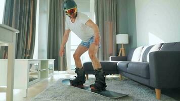 Fun video. Man in shorts and a T-shirt depicts snowboarding on a carpet in a cozy room. Waiting for a snowy winter video