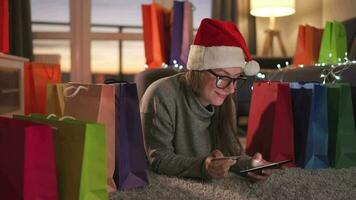 Happy woman with glasses wearing a santa claus hat is lying on the carpet and makes an online purchase using a credit card video