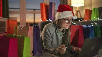 contento mujer con lentes vistiendo un Papa Noel claus sombrero es acostado en el alfombra y hace un en línea compra utilizando un crédito tarjeta video