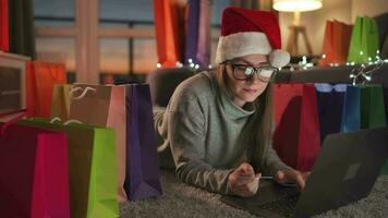 contento mujer con lentes vistiendo un Papa Noel claus sombrero es acostado en el alfombra y hace un en línea compra utilizando un crédito tarjeta video