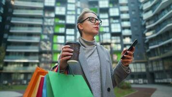 mujer caminando en un negocio distrito sostiene para llevar café compras pantalones y usos teléfono inteligente video